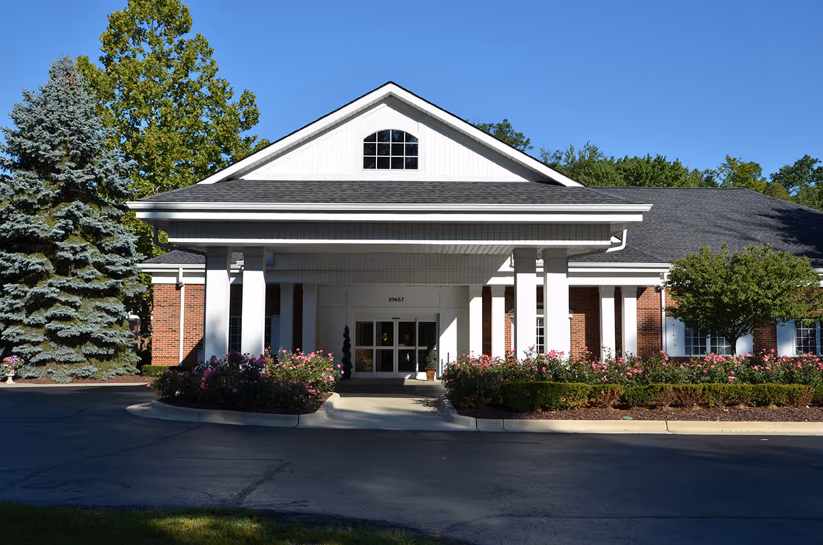 Front exterior view of Woodhaven Retirement Community building with a covered entrance supported by white columns, surrounded by landscaped bushes and trees under a clear blue sky.