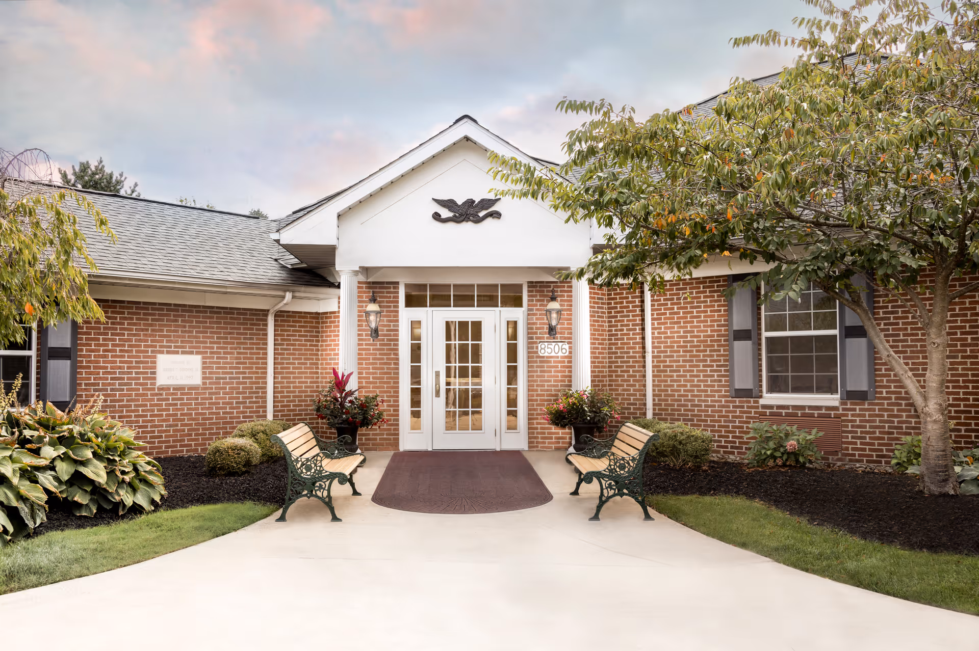 Front entrance of a brick building with white double doors, two benches on either side of the walkway, potted plants, and trees surrounding the entrance under a partly cloudy sky.