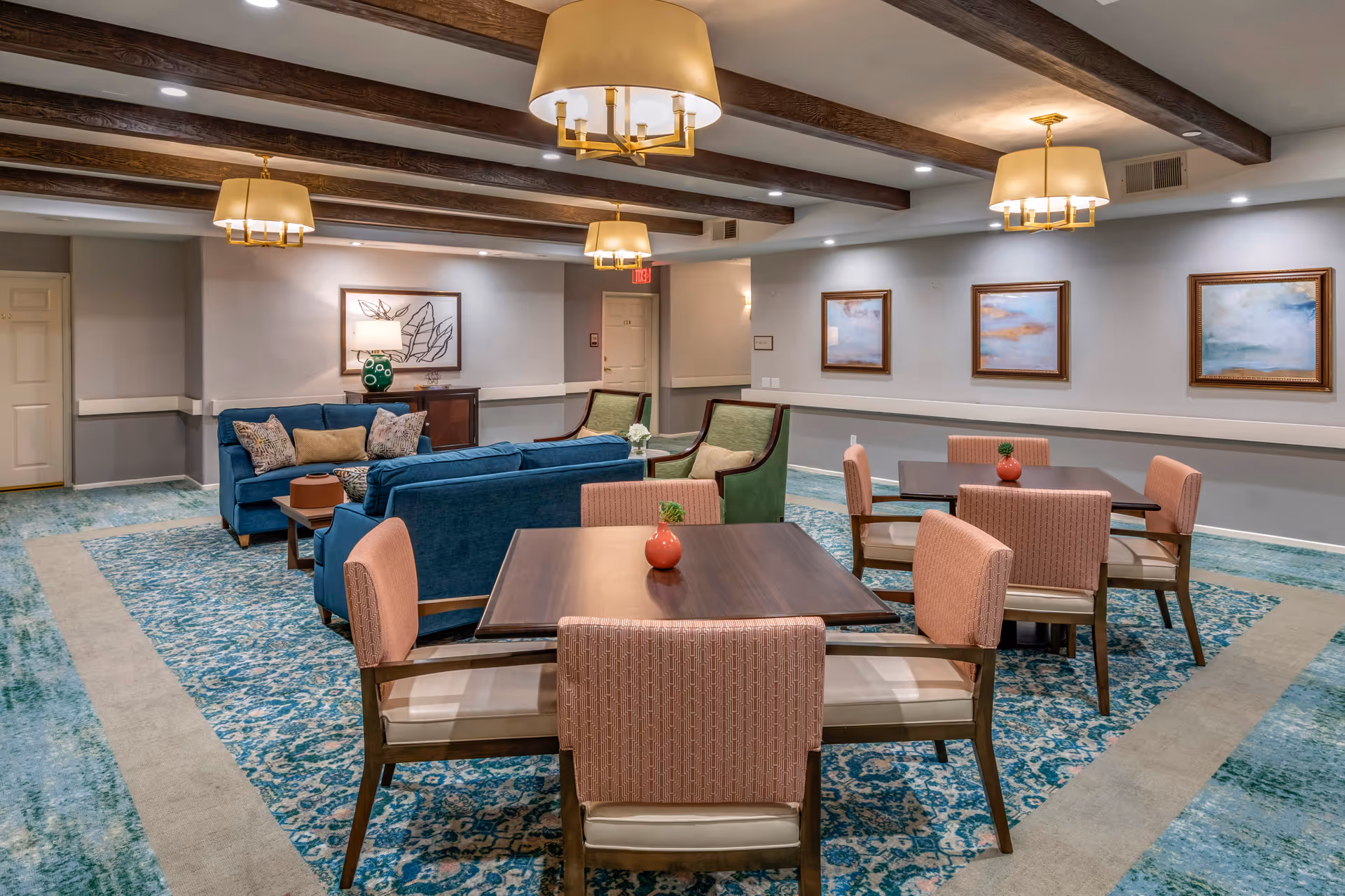 A cozy common area in a senior living facility featuring two blue sofas, two green armchairs, and two wooden tables each surrounded by four pink upholstered chairs. The room has a patterned blue and beige carpet, wooden ceiling beams, and three framed paintings on the wall. Warm ceiling lights illuminate the space.