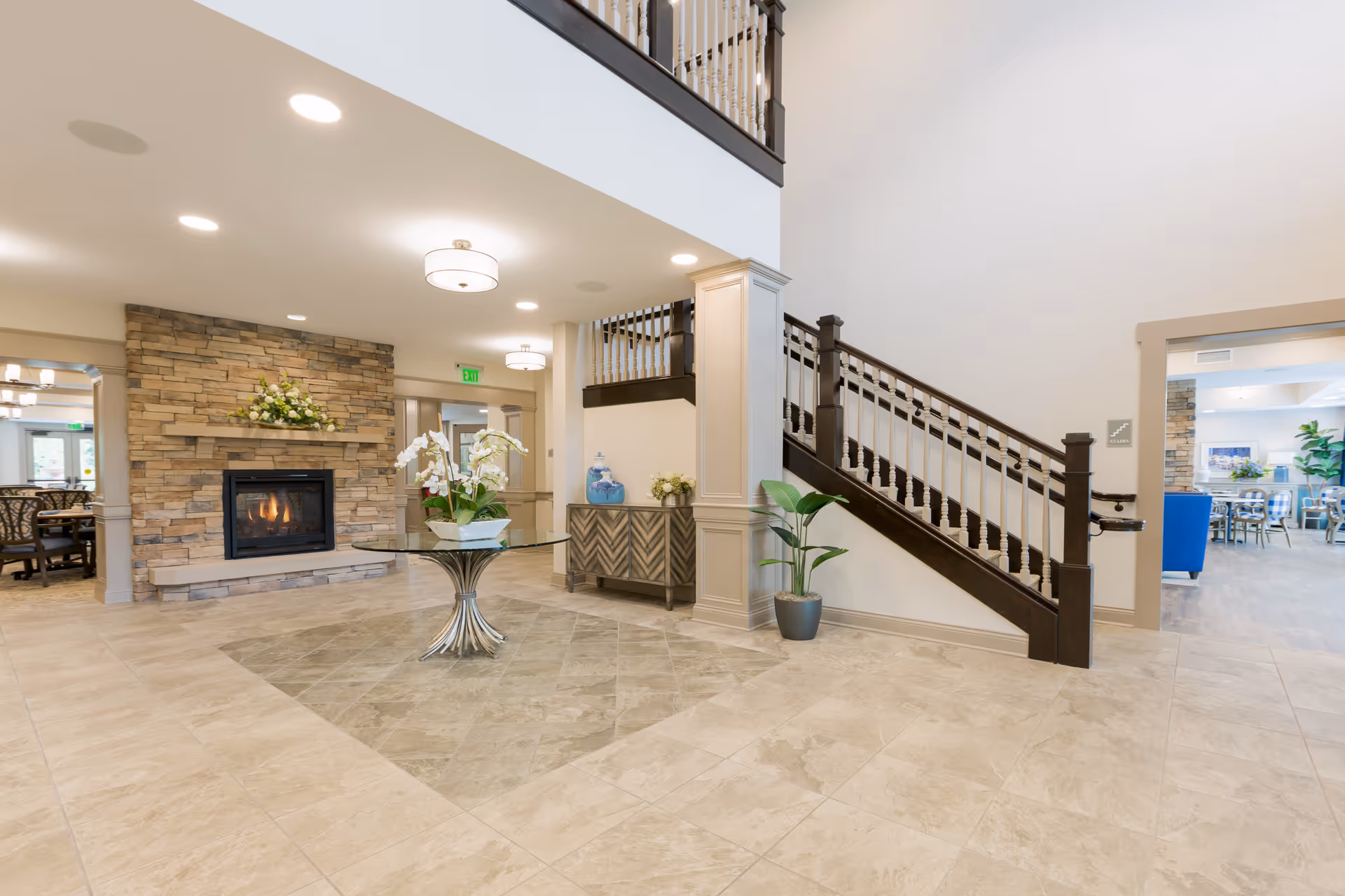Bright and spacious interior of Mason Assisted Living & Memory Care featuring a tiled floor, a stone fireplace with a floral arrangement on the mantel, a round glass table with white orchids in the center, a staircase with dark wood railing, and a sideboard with decorative items. Adjacent rooms with dining tables and chairs are visible.