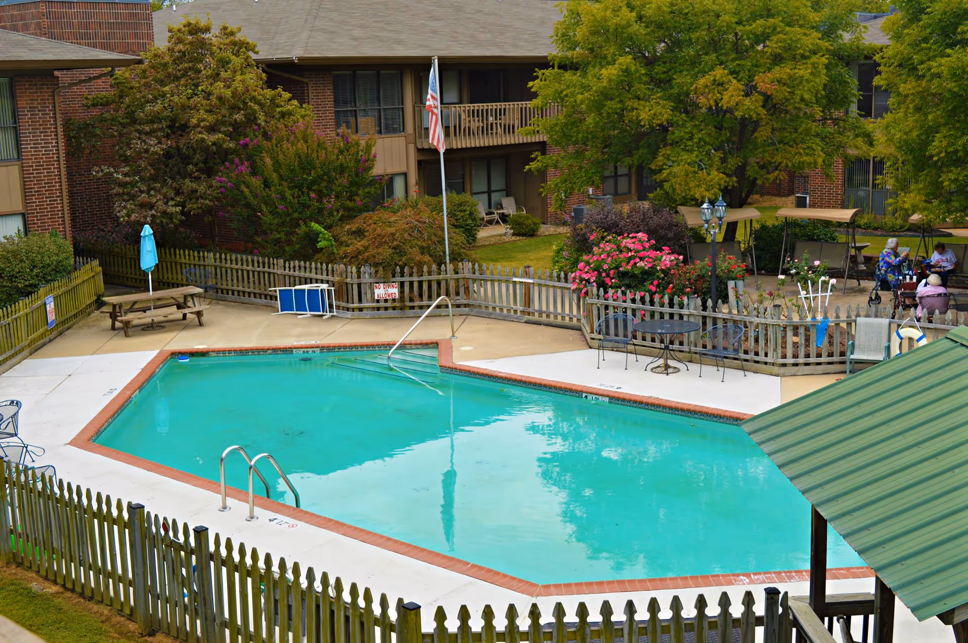 A fenced outdoor swimming pool and surrounding patio with tables, chairs, flowers and apartment balconies in the background.