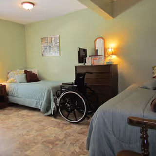 A room with two single beds covered in light blue bedding, a wheelchair positioned between the beds, a wooden dresser with a mirror and a lamp on top, and a collage of photos on the light green wall.