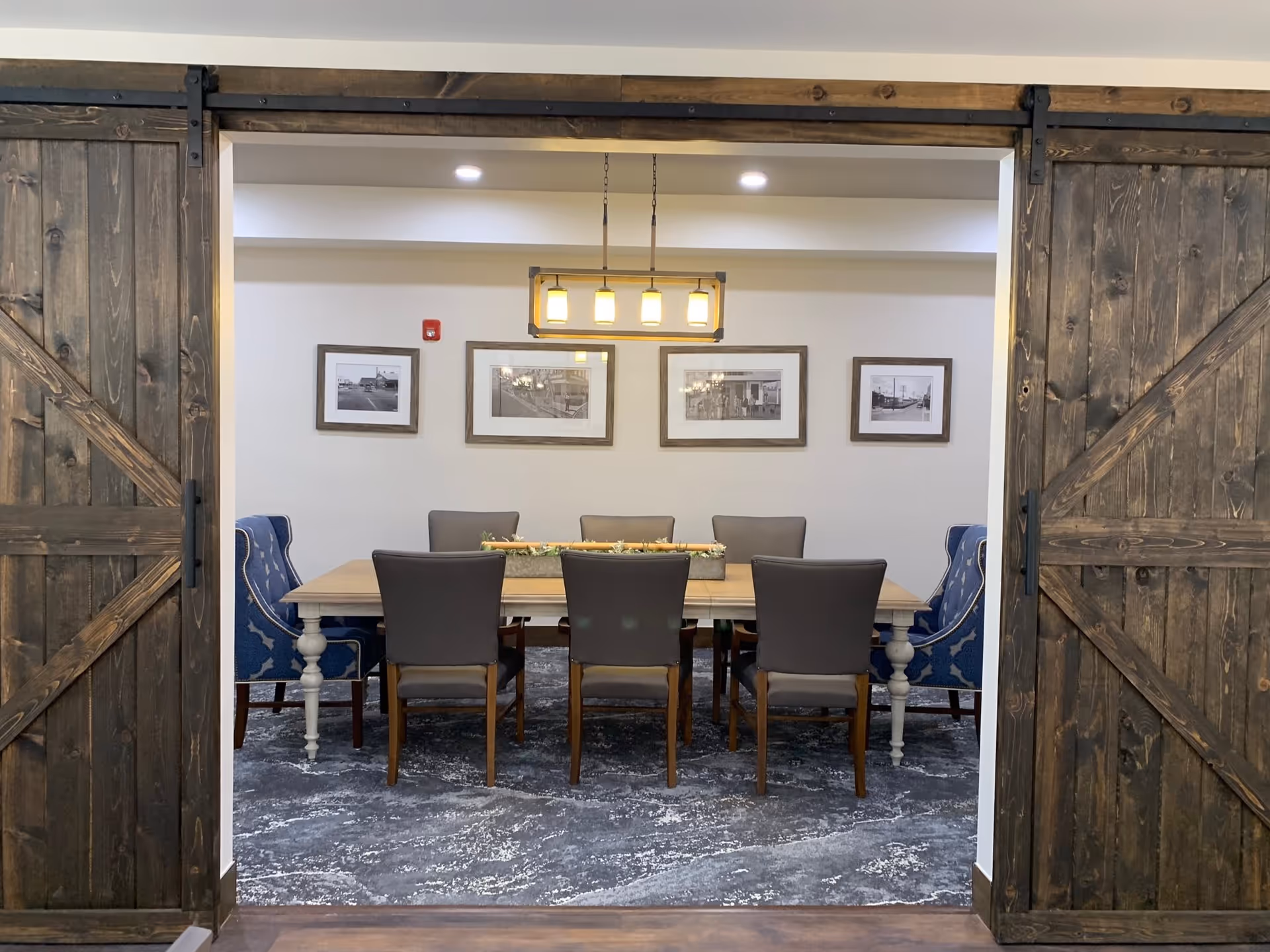 A dining room with a long wooden table surrounded by eight chairs, two of which have blue upholstery with white patterns. The room is viewed through a large doorway framed by rustic sliding barn doors. Above the table hangs a rectangular light fixture with four lights. Four framed black and white photographs are displayed on the wall behind the table.