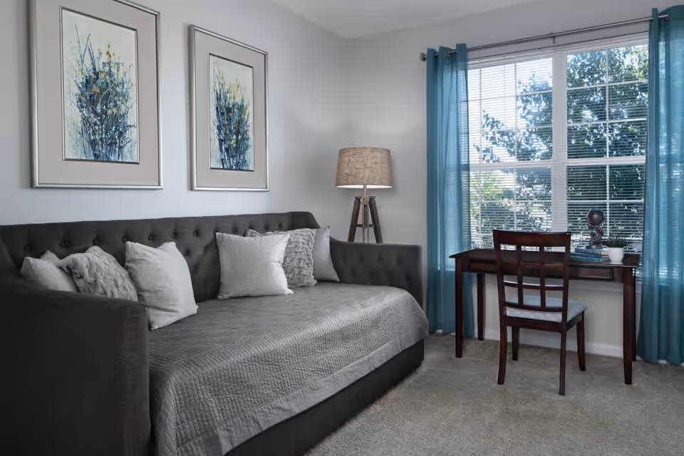 A cozy living room with a dark gray tufted sofa covered with a gray quilt and several light-colored pillows. Two framed abstract floral paintings hang on the light gray wall above the sofa. To the right, there is a wooden desk with a matching chair placed in front of a large window with blue curtains. A tripod floor lamp with a beige shade stands between the sofa and the desk.