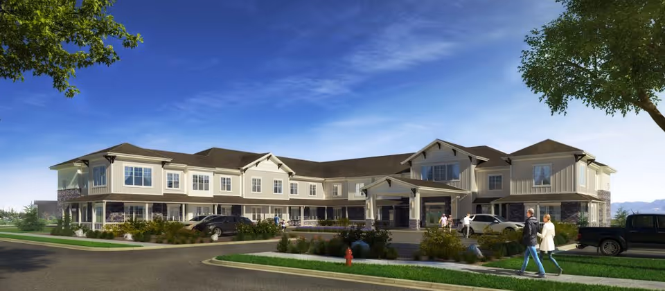 Exterior view of a two-story senior living facility building with beige siding and stone accents, surrounded by landscaped greenery and a clear blue sky. Several people are walking near the entrance, and cars are parked in front of the building.