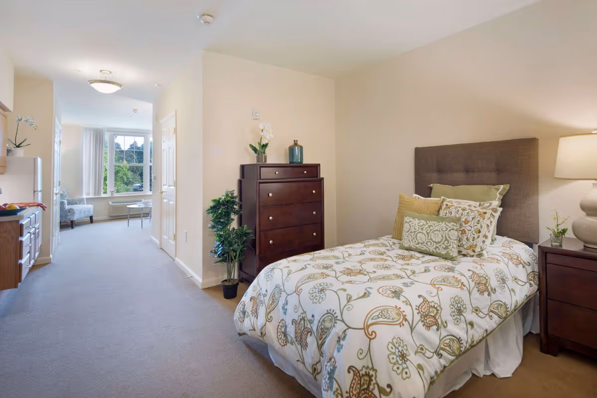 A bright and spacious bedroom in a senior living facility featuring a neatly made bed with patterned bedding and multiple pillows. Next to the bed is a wooden nightstand with a lamp and a small plant. Across from the bed is a tall wooden dresser with decorative items on top and a potted plant beside it. The room opens into a hallway leading to a sitting area with a large window letting in natural light.