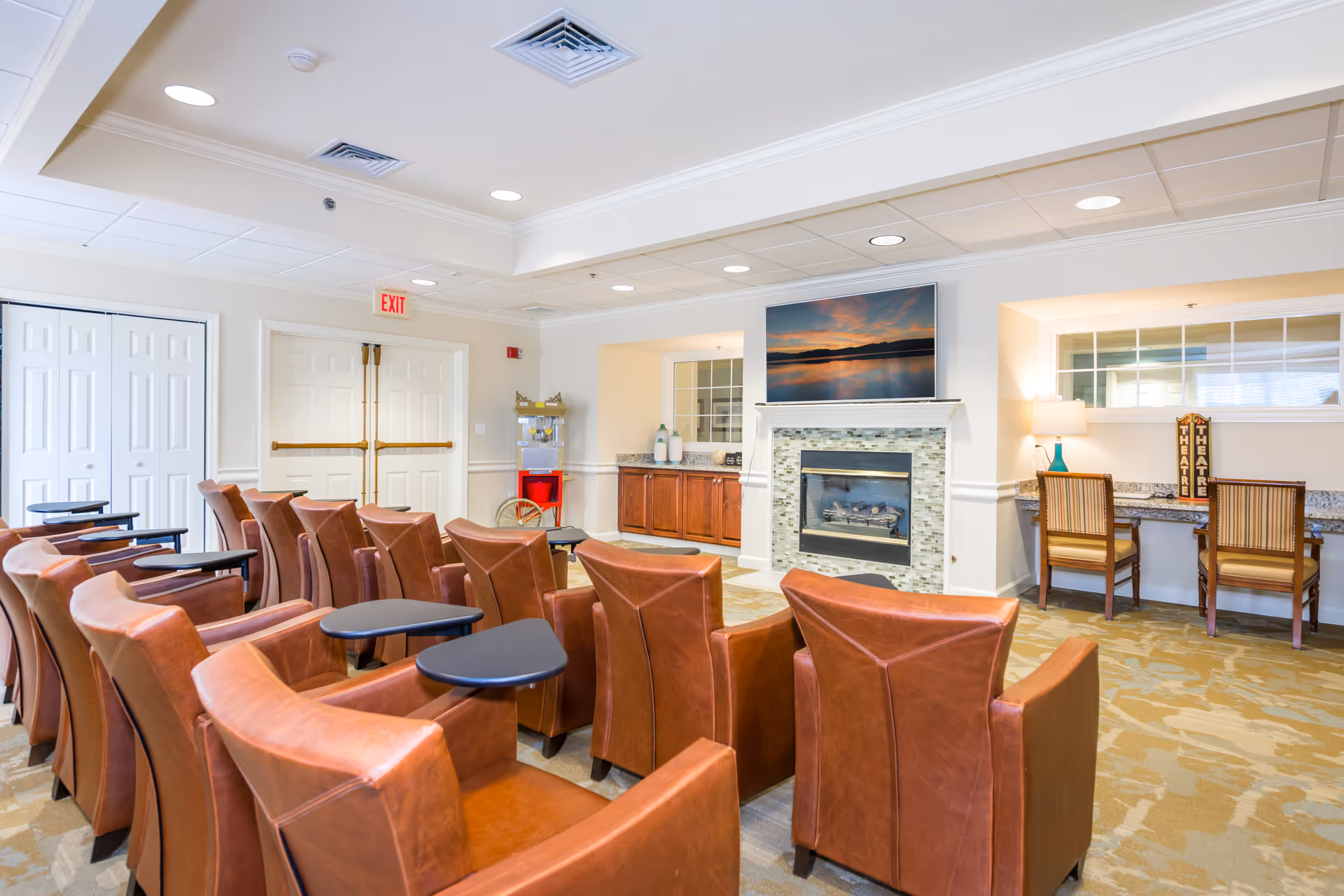 A cozy common area with rows of brown leather chairs equipped with small black foldable tables facing a fireplace with a mounted TV above it displaying a scenic sunset over water. To the right, there is a countertop with two wooden chairs, a lamp, and decorative items. The room has a light-colored carpet, white walls, and a ceiling with recessed lighting.