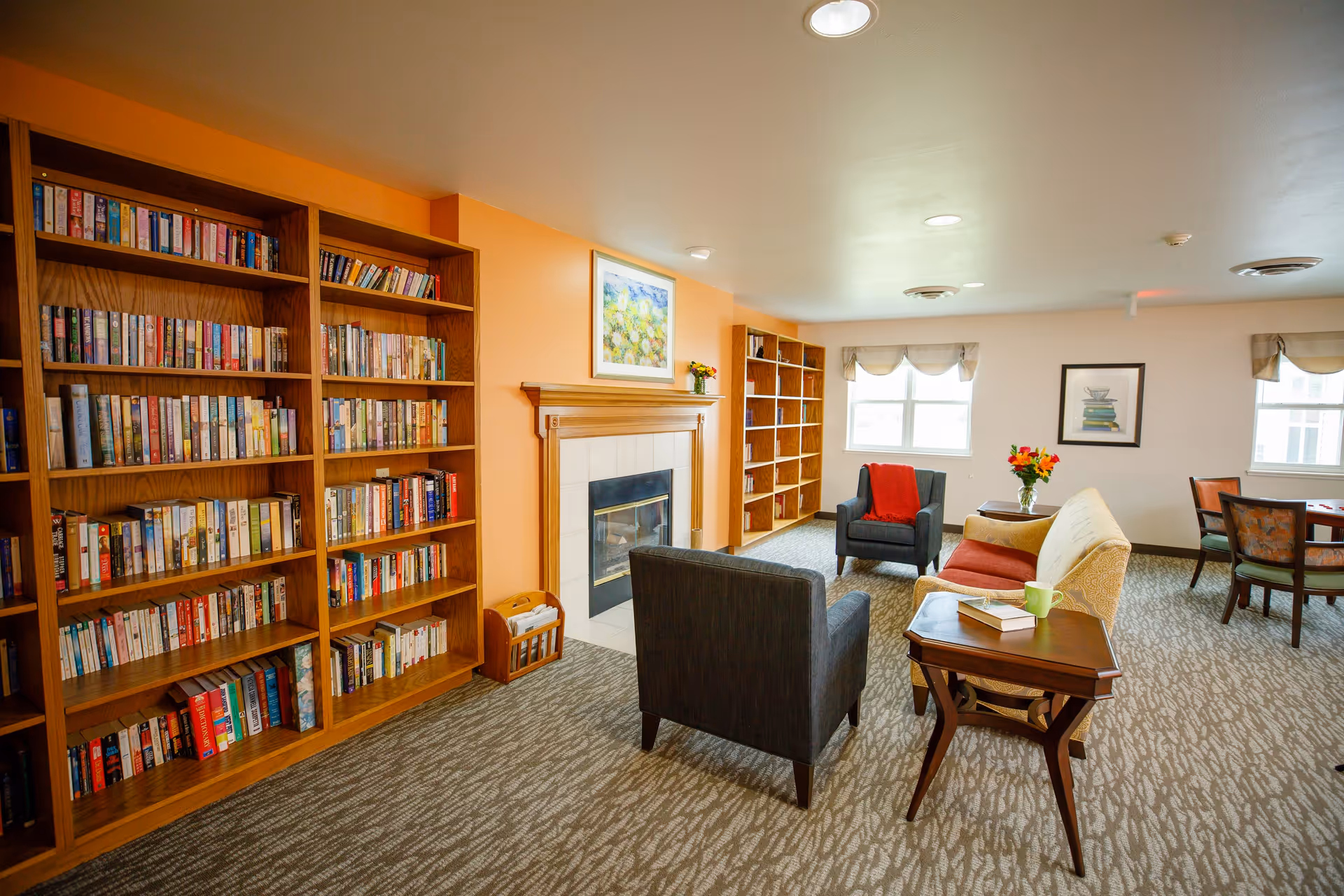 A cozy living room area with wooden bookshelves filled with books along the left wall, a fireplace with a wooden mantle and a framed painting above it, two armchairs and a sofa arranged around a small wooden side table with a book and a green mug on it. There are windows with beige valances letting in natural light, a small table with a vase of flowers, and a dining table with chairs in the background.