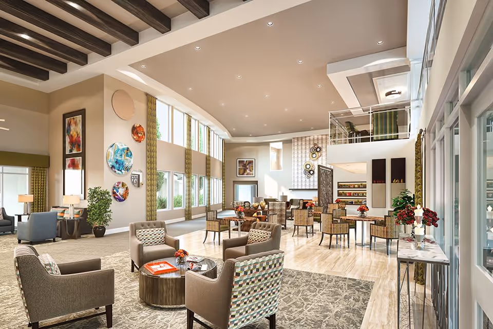 A spacious and well-lit senior living facility common area with high ceilings and large windows. The room features multiple seating arrangements including armchairs around small tables, decorative wall art, plants, and a mezzanine level with additional seating. The space is decorated with modern furnishings and warm neutral tones.