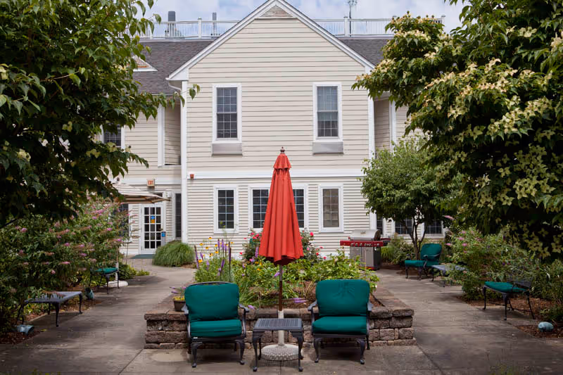 Outdoor patio area with two green cushioned chairs and a small table between them, centered in front of a red closed umbrella. The patio is surrounded by greenery, trees, and flowering plants, with a beige two-story building in the background.