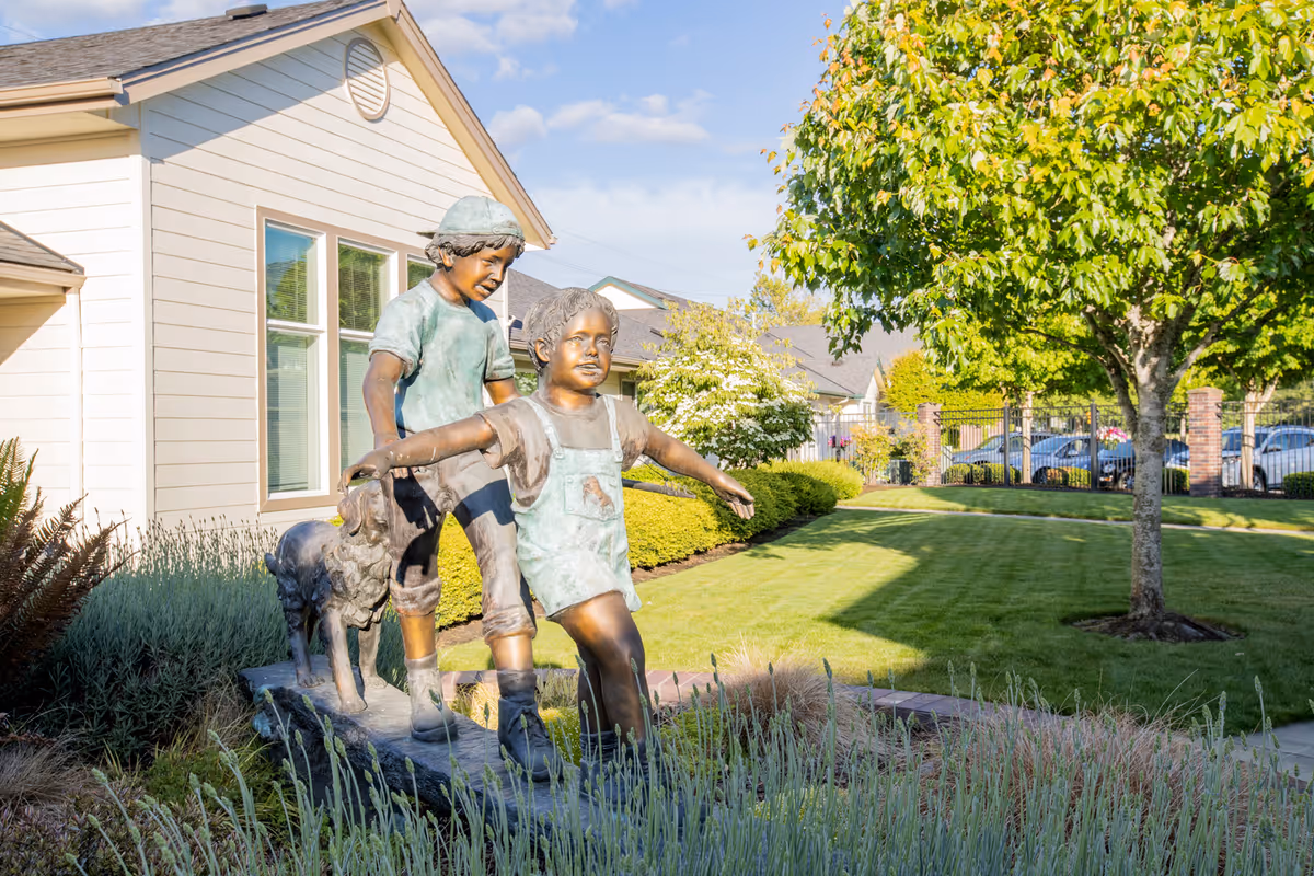 Bronze statue of two children and a dog on a landscaped lawn in front of a light-colored building.