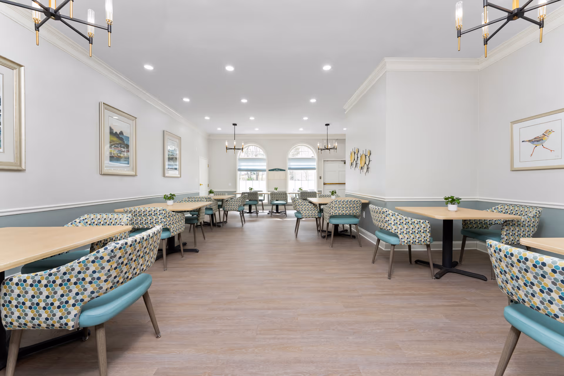 A bright and spacious dining room with multiple tables and patterned chairs arranged neatly. The room features light-colored walls with framed artwork, large arched windows letting in natural light, and modern chandeliers hanging from the ceiling. The floor is wooden, and small potted plants are placed on the tables.