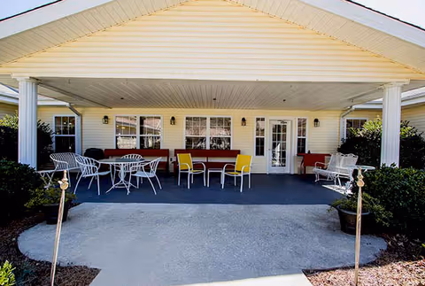 Covered outdoor patio area at Langston Square with white columns, several white metal chairs and tables, red cushioned benches, and yellow chairs. The patio is in front of a building with cream-colored siding and multiple windows and doors.