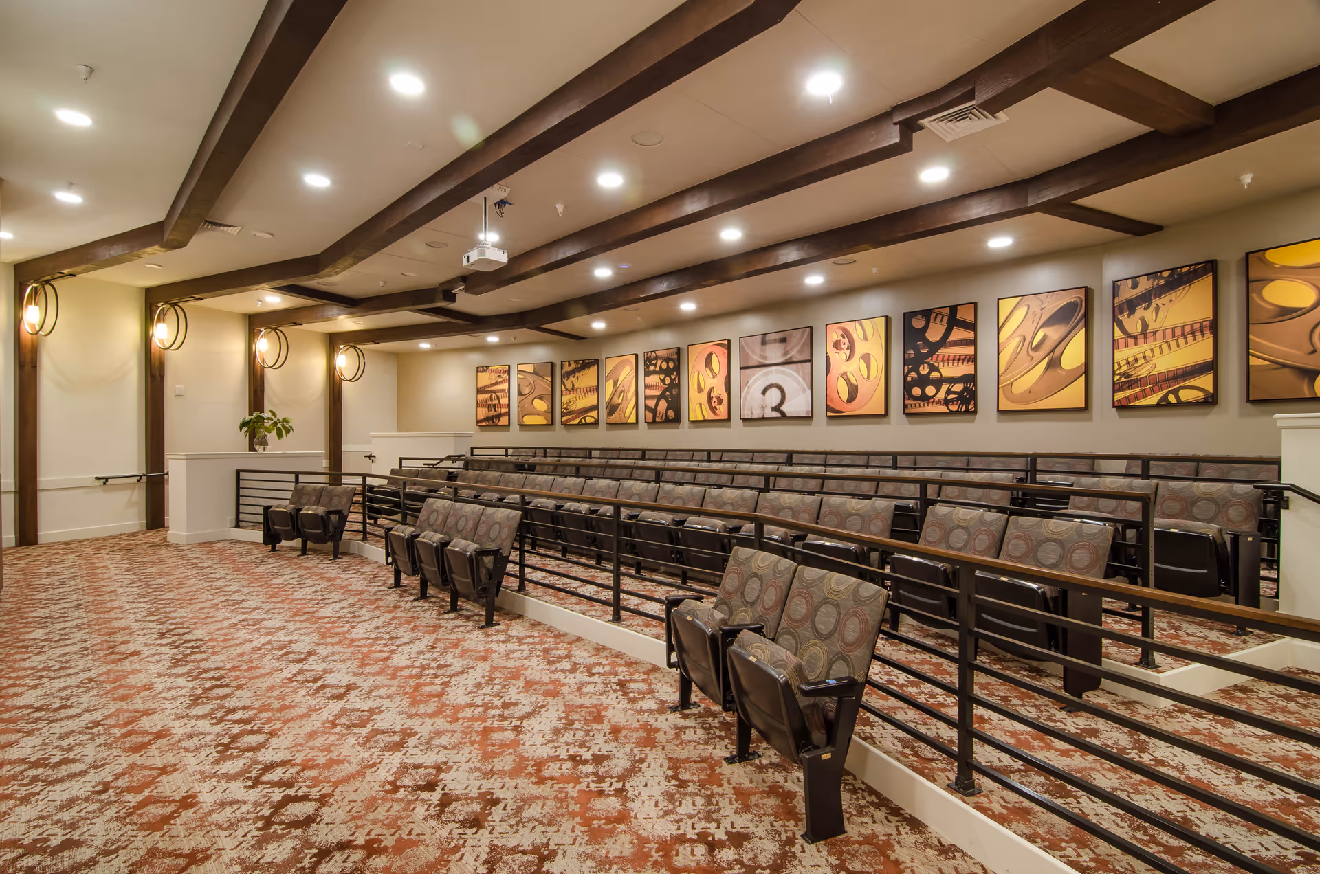 Interior view of a small auditorium or theater room with tiered seating featuring cushioned chairs with patterned upholstery. The room has a patterned carpet, wooden ceiling beams, recessed lighting, and framed abstract artwork on the walls.