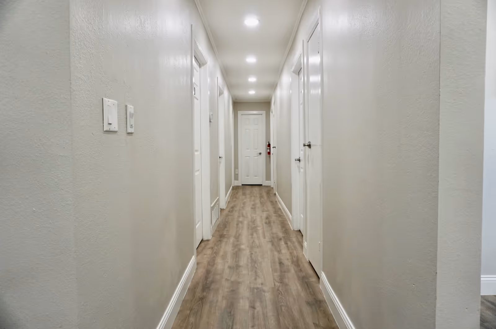 Long narrow interior hallway with wood-look flooring, recessed ceiling lights and multiple white doors along both sides.