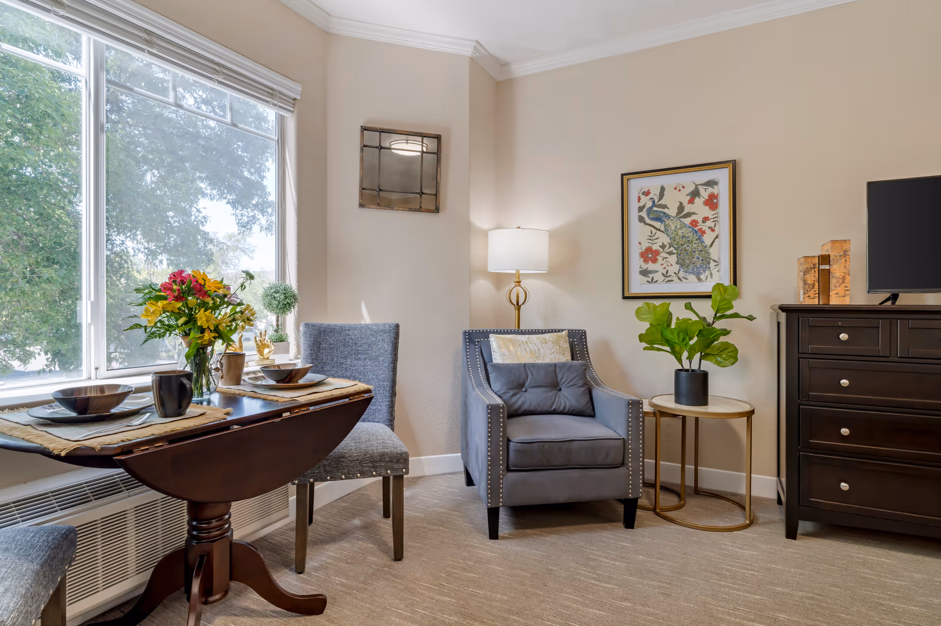 Sunlit sitting area with an upholstered armchair, side table, dresser and a small dining table set by a large window.