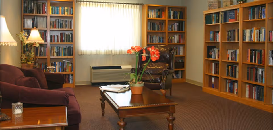 Cozy common room with bookshelves lining the walls, armchairs, a coffee table with a potted plant, and a curtained window.