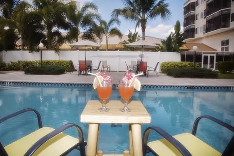 Two tropical cocktails on a small table by a swimming pool with lounge chairs, palm trees and a building in the background.