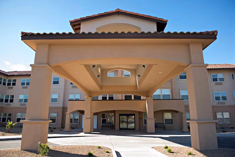 Front exterior view of Joshua Springs Senior Living facility showing a large covered entrance with columns, beige stucco walls, multiple windows, and a tiled roof under a clear blue sky.