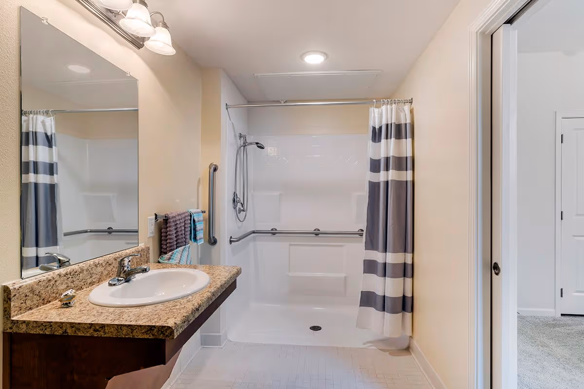 A clean and accessible bathroom featuring a wall-mounted sink with a granite countertop, a large mirror above the sink, and a walk-in shower with grab bars and a striped shower curtain. The bathroom has beige walls and a white tiled floor. A towel rack with two towels is mounted on the wall next to the shower.