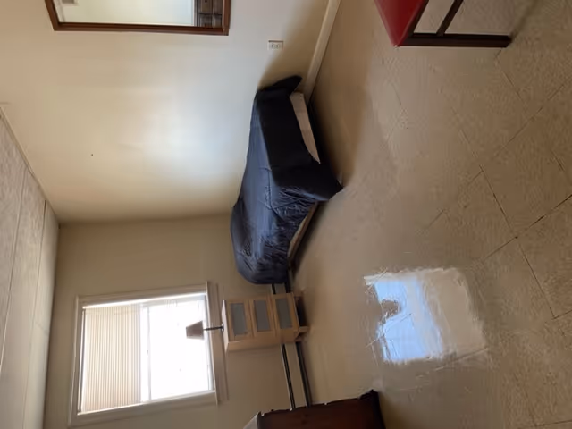 A simple bedroom with a single bed covered with a dark blanket against a beige wall. There is a small wooden nightstand with a lamp next to the bed under a window with closed blinds. A wooden dresser is partially visible on the left side, and a mirror hangs on the right wall. The floor is tiled and shiny, reflecting some light from the window.