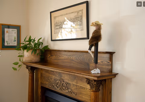 A wooden mantelpiece with decorative carvings, featuring a potted green plant on the left and a small figurine wearing a hat and fur coat on the right. Above the mantel hangs a framed black and white artwork. The wall is painted a light color.