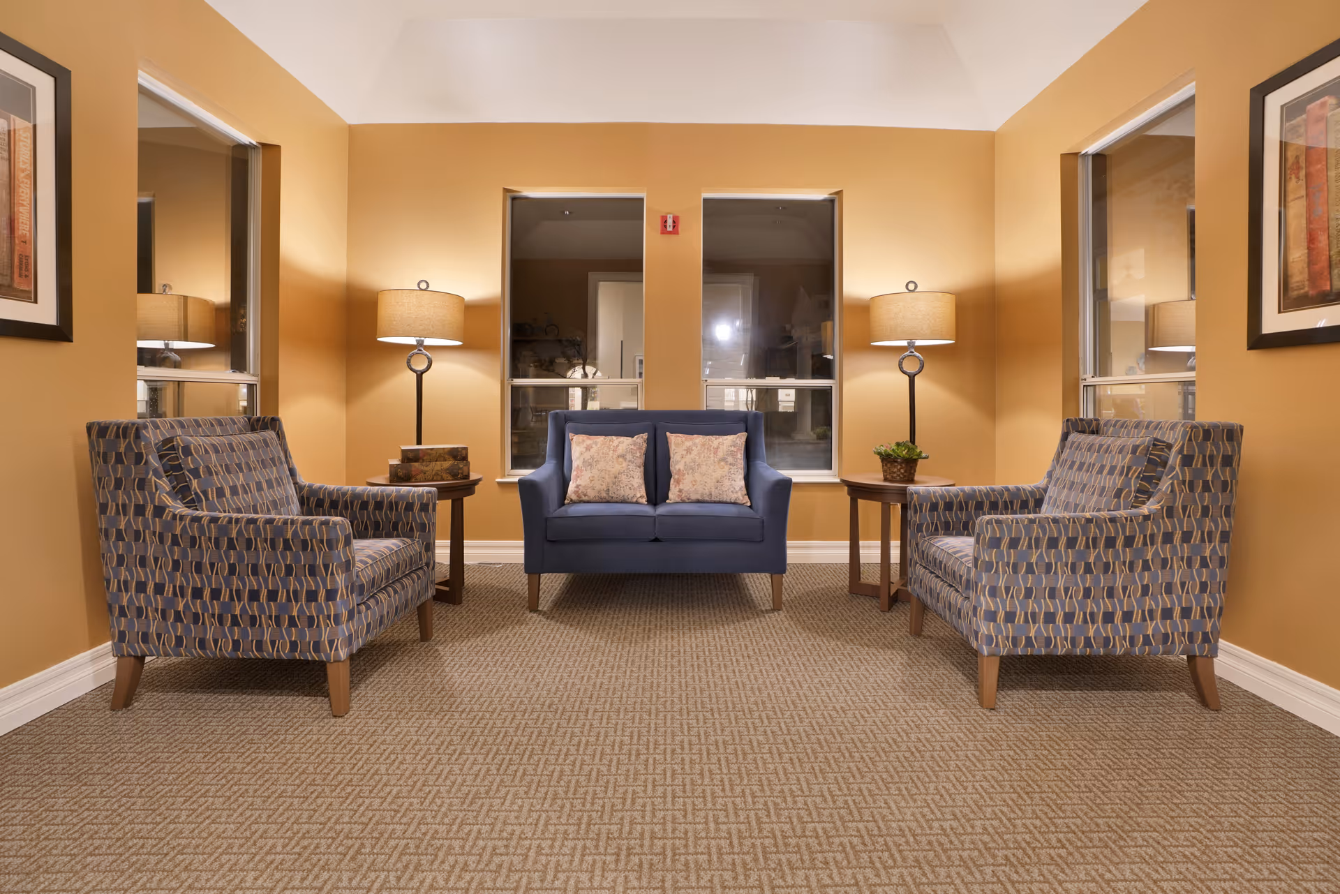 A cozy sitting area with a blue loveseat with two floral pillows in the center, flanked by two patterned armchairs on either side. Each armchair has a wooden side table next to it with a lamp. The walls are painted a warm beige color, and there are three windows behind the loveseat. The floor is carpeted with a textured pattern, and framed artwork is visible on the walls.