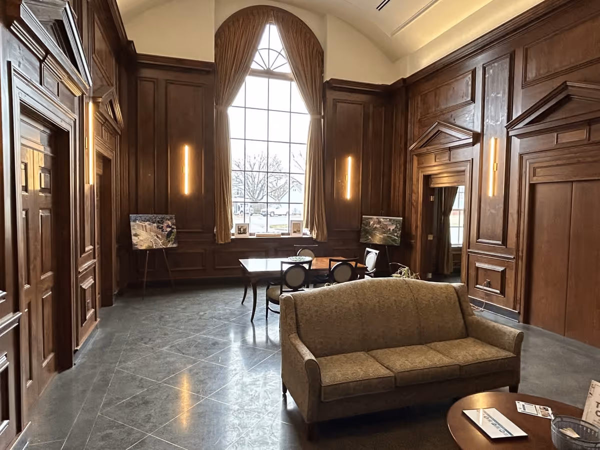 Wood-paneled communal lounge with a tall arched window, a sofa in the foreground and a table with chairs near the window.