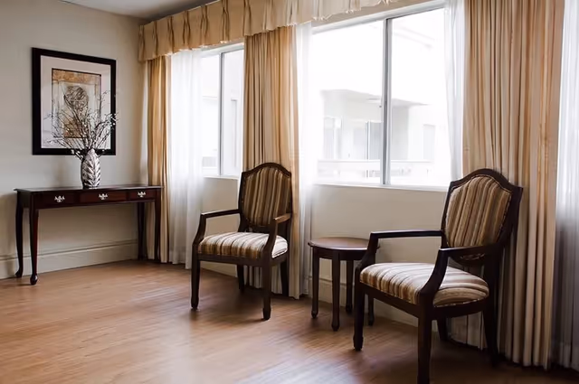 A bright room with two wooden armchairs with striped cushions positioned near large windows with beige curtains. A small round wooden table is placed between the chairs. On the left side, there is a dark wooden console table with three drawers and a decorative vase with branches on top. A framed artwork hangs on the wall above the console table. The floor is wooden.