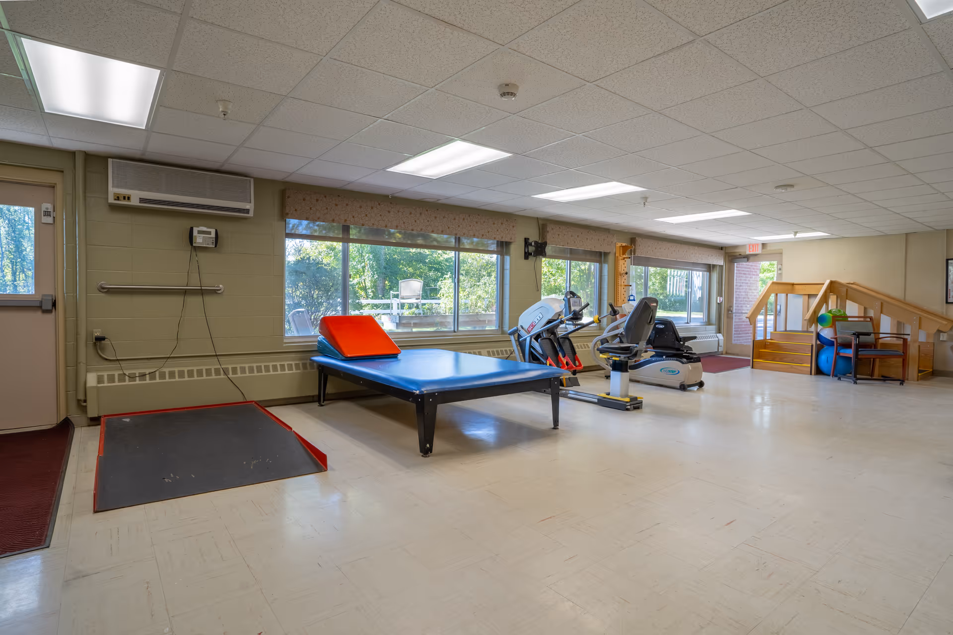 A spacious physical therapy or exercise room with large windows letting in natural light. The room contains exercise equipment including a recumbent bike, a treadmill, a padded therapy table with a red wedge cushion, a ramp, and a set of stairs with handrails. There are also exercise balls and chairs near the stairs. The walls are light-colored, and the floor is a light vinyl tile.