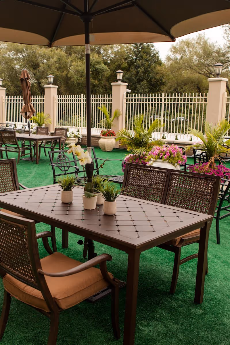 Outdoor patio area with several brown metal tables and chairs on green artificial turf. Tables have small potted plants and flowers on them. Large umbrellas provide shade, and there are decorative plants and flowers around the fenced perimeter with trees in the background.