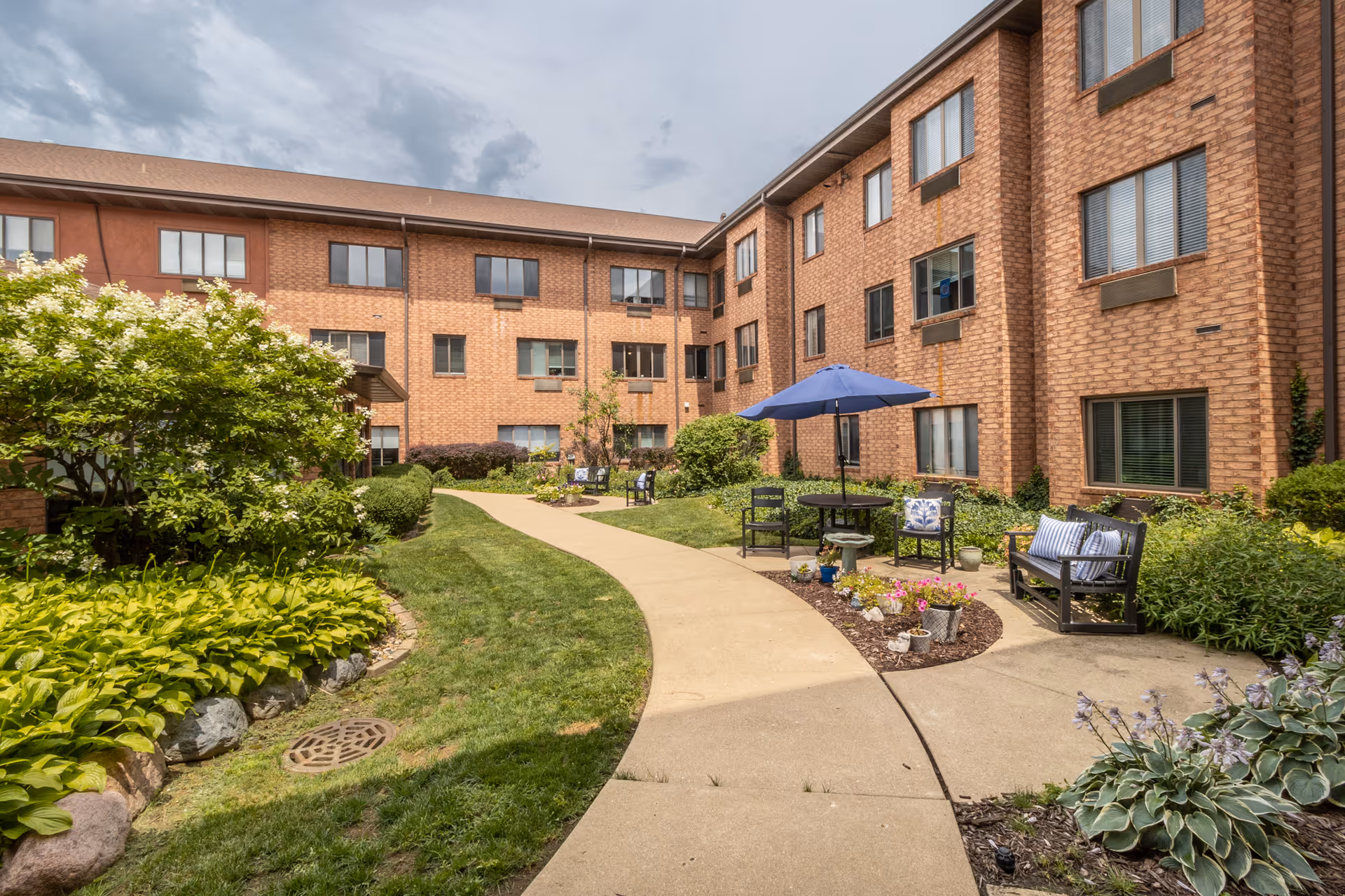 Brick three-story senior living building surrounding a landscaped courtyard with a curved walkway, benches, potted flowers and a blue patio umbrella.