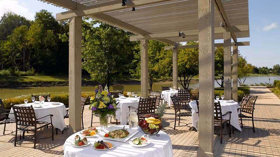 Outdoor dining area with several round tables covered with white tablecloths and set with plates, glasses, and cutlery under a wooden pergola. There are metal chairs around the tables and a centerpiece of flowers on one table. The area overlooks a body of water with trees and greenery in the background under a clear sky.