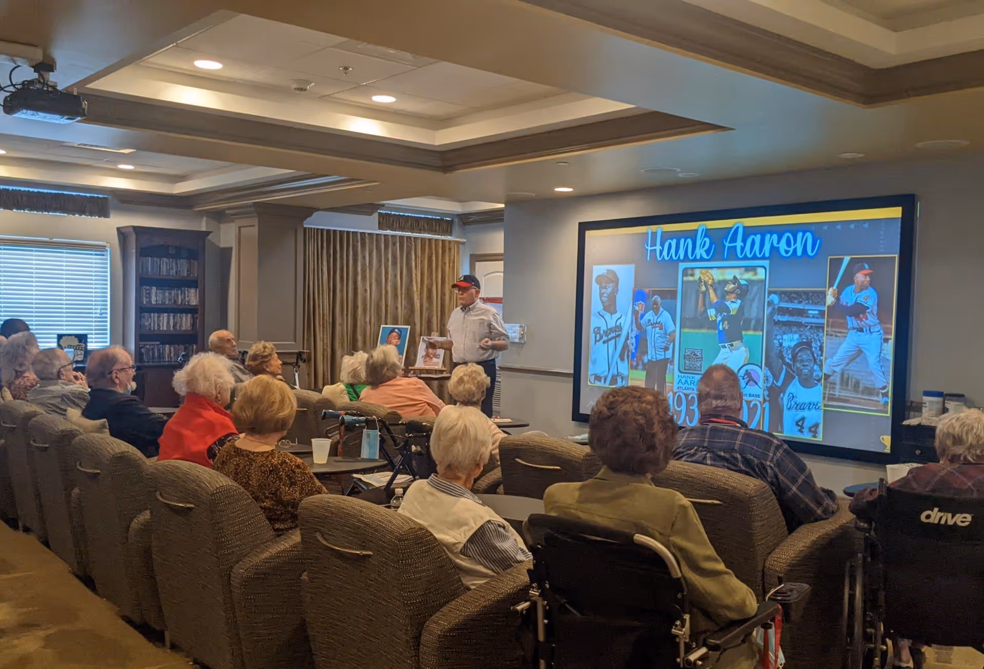 A group of elderly people seated in a common room watching a presentation about Hank Aaron on a large screen. The presenter stands near the screen, and some attendees are in wheelchairs. The room has comfortable chairs, a bookshelf, and windows with blinds.