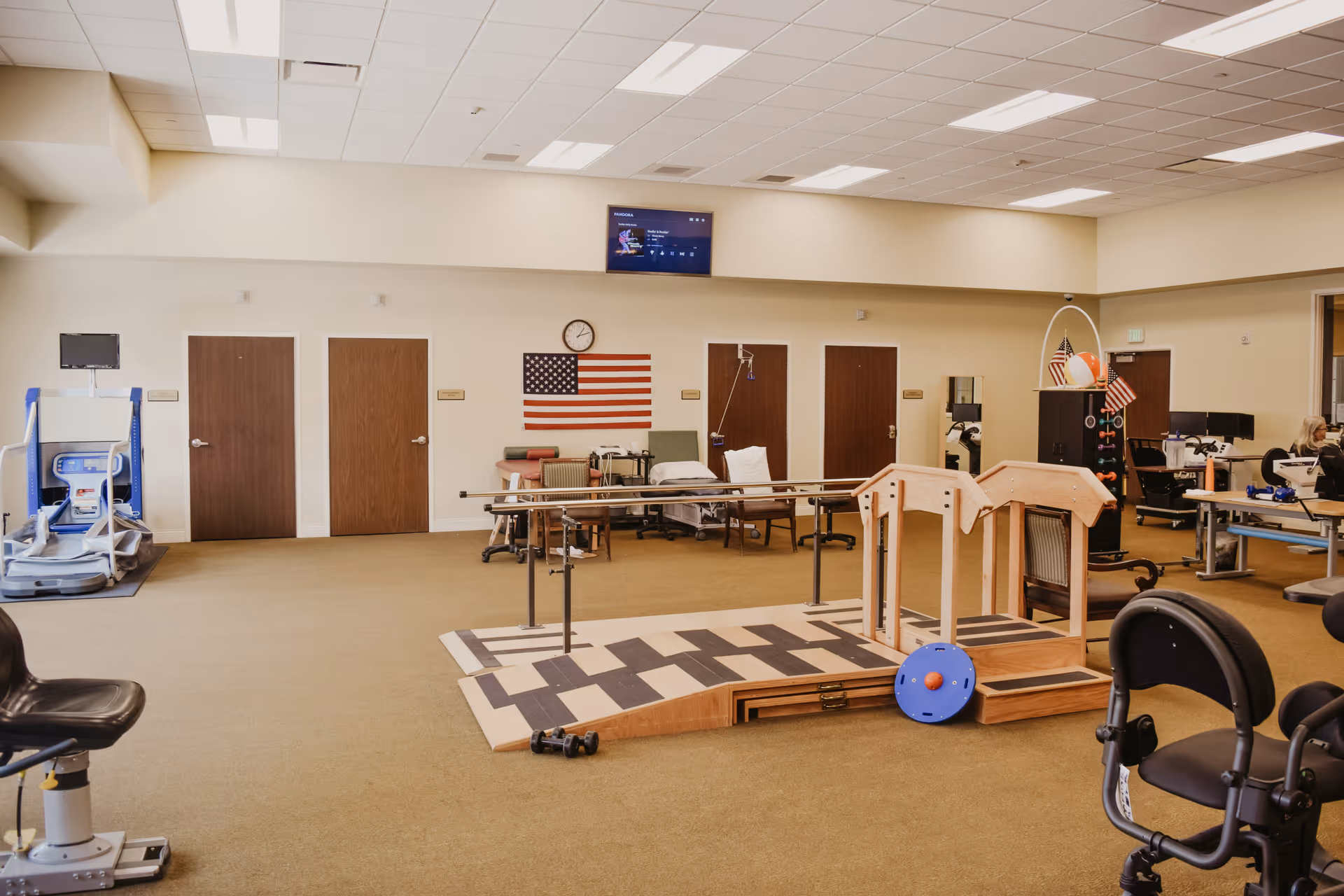 A spacious rehabilitation therapy room with exercise equipment, parallel bars for walking practice, chairs, and therapy beds. The room has beige walls, a clock, an American flag on the wall, and a television mounted above. Several closed wooden doors are visible along the back wall.