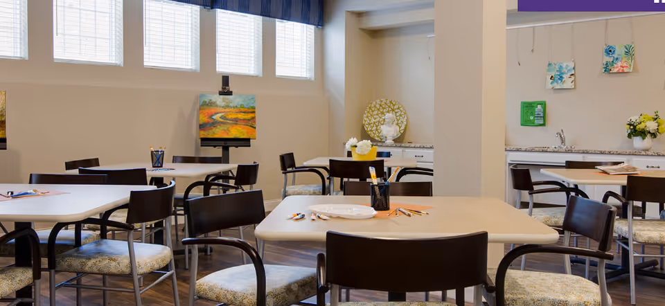 A bright, well-lit room with multiple tables and chairs arranged for group activities or dining. The tables have art supplies like colored pencils and paper. The walls feature colorful paintings and decorative items, with windows near the ceiling allowing natural light in.