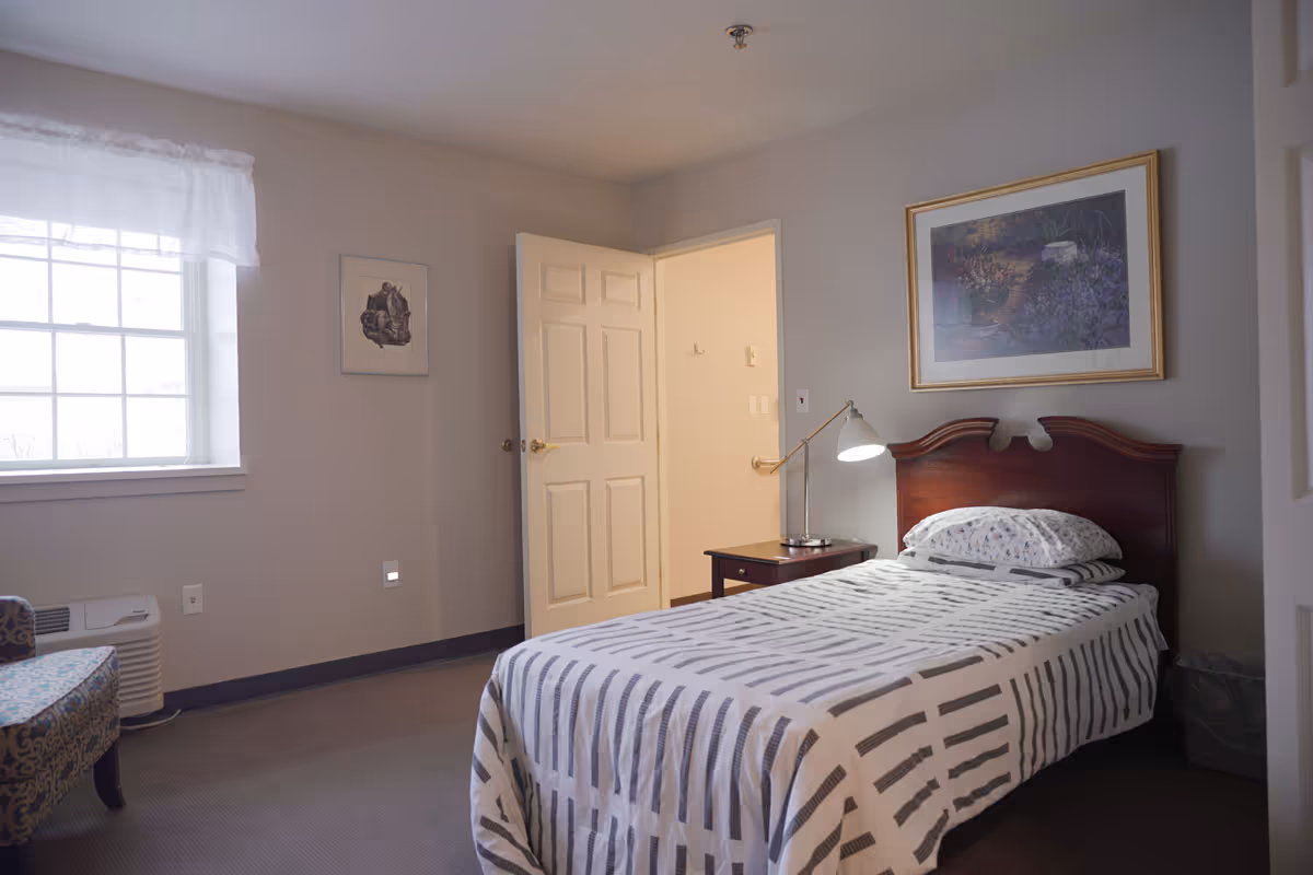 A simple bedroom with a single bed covered in a white and gray striped bedspread, a wooden headboard, a bedside table with a lamp, a framed picture on the wall above the bed, a window with sheer white curtains, and an open door leading to another room.