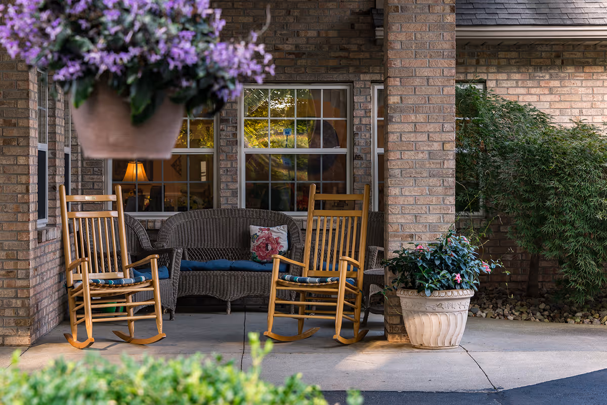Covered brick porch with two wooden rocking chairs, a wicker loveseat, and potted plants in front of windows.