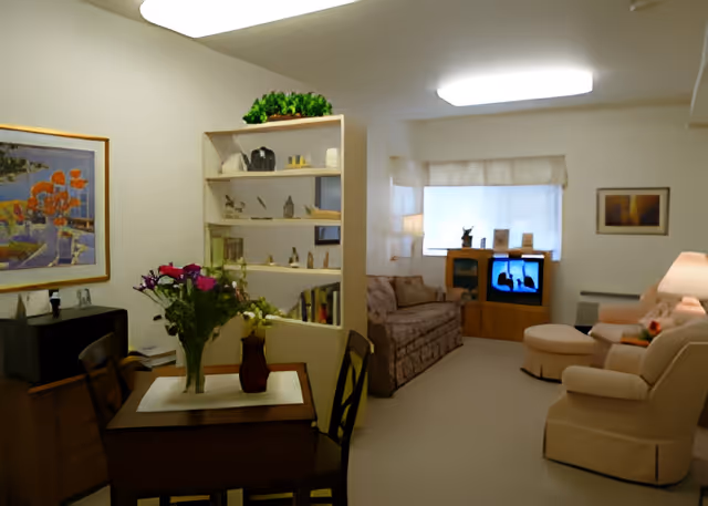 A cozy living room and dining area in a senior living facility. The dining area has a small wooden table with a vase of flowers and chairs around it. A white bookshelf with decorative items separates the dining area from the living room. The living room features a patterned sofa, two beige armchairs with an ottoman, a wooden TV stand with a television, and framed artwork on the walls. Soft lighting creates a warm and inviting atmosphere.