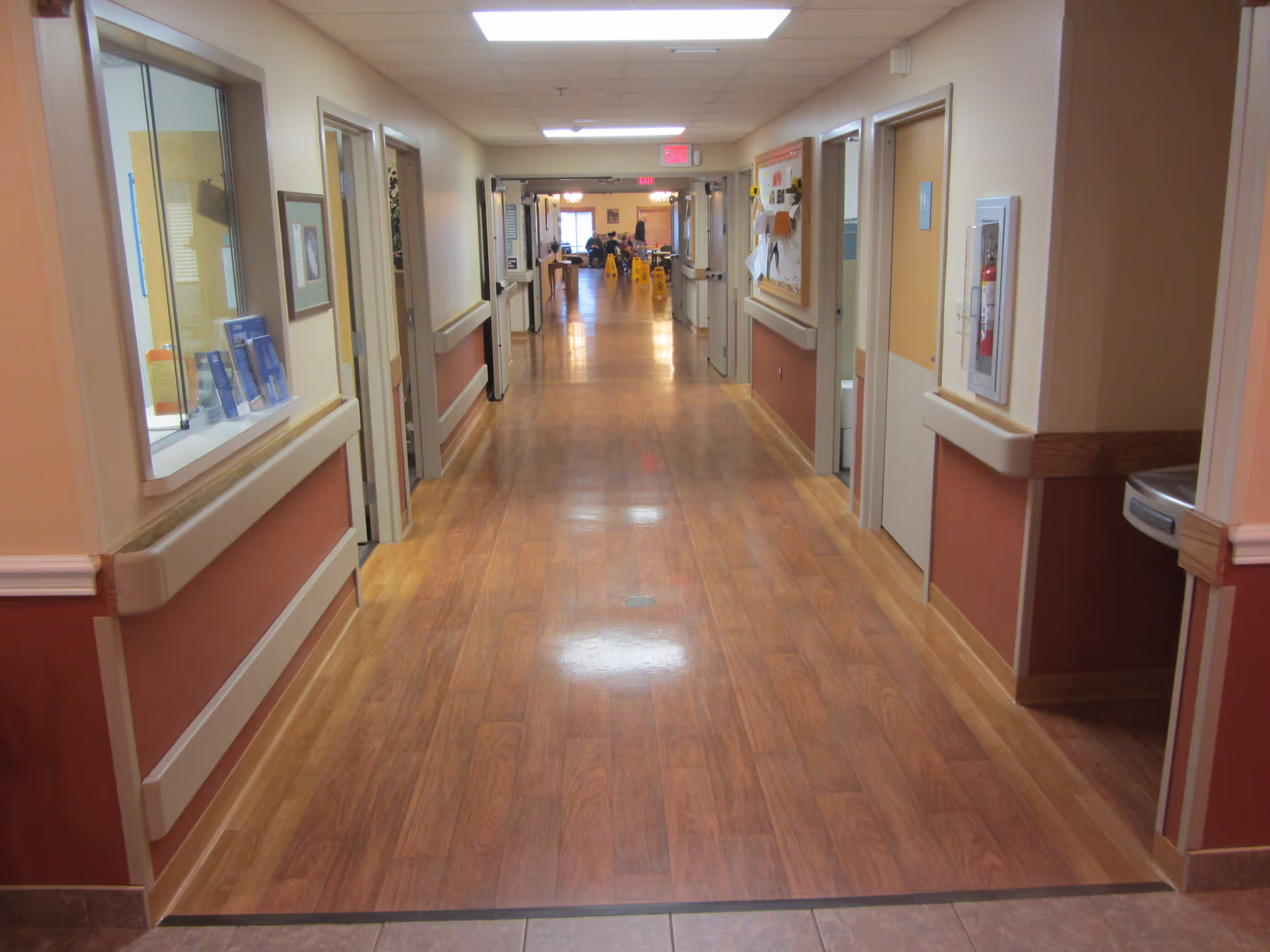 A long, clean hallway in a healthcare facility with wood-patterned flooring, beige walls with maroon lower panels, handrails on both sides, several doors, a bulletin board, a fire extinguisher, and a water fountain. The hallway is well-lit with ceiling lights and leads to a room with tables and chairs at the far end.