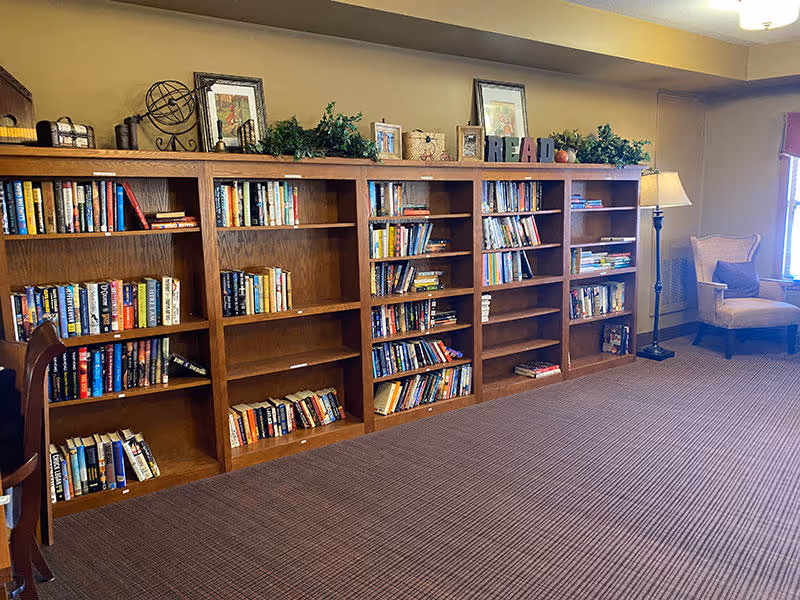 A cozy room with a long wooden bookshelf filled with books, decorative items on top including framed pictures, greenery, and letters spelling READ. To the right, there is a floor lamp and a cushioned armchair near a window with natural light.
