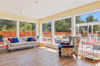 A bright living room with large windows showing an outdoor patio area. The room features a light-colored sofa with colorful pillows, a patterned armchair, a small side table with a plant, and a rolling cart with glass shelves. The floor is wooden, and sunlight fills the space.