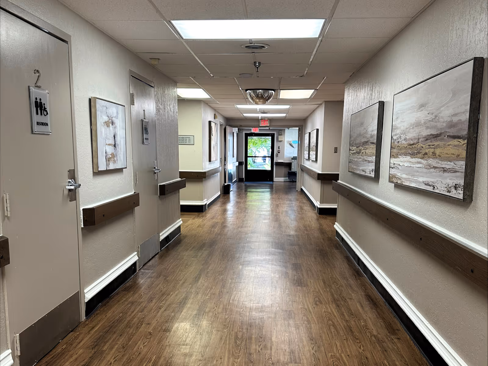 Interior corridor of a senior living facility with wood floors, handrails, doors and framed artwork leading to an exit door.