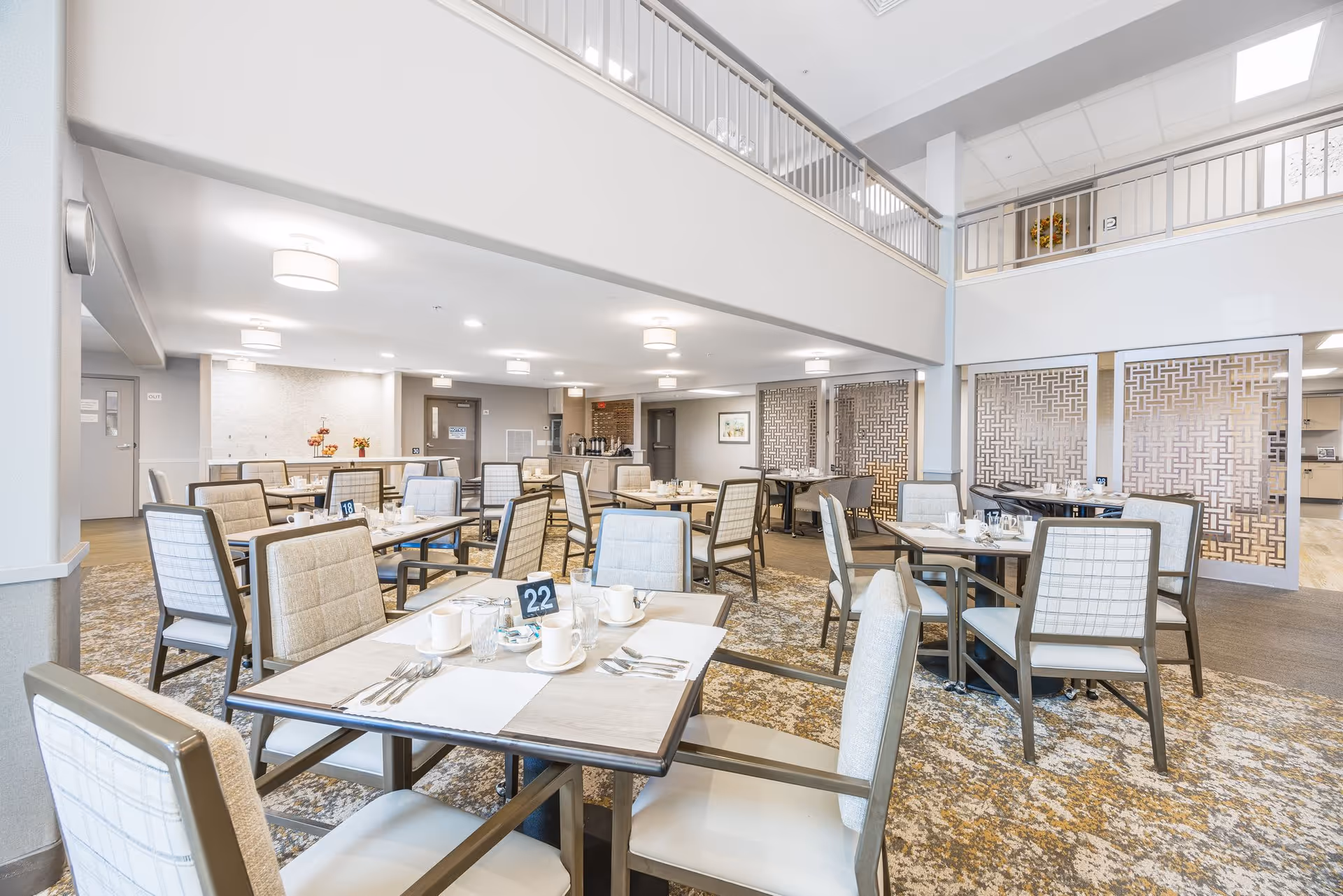 Bright and spacious dining room with multiple tables and chairs arranged neatly. Each table is set with white cups, glasses, and silverware. The room features a high ceiling with a second-floor balcony overlooking the area. Decorative partition panels separate the dining area from the kitchen in the background.