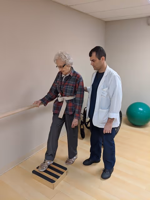 An elderly woman is performing a physical therapy exercise by stepping onto a small wooden platform with black stripes while holding onto a handrail. A healthcare professional in a white coat is standing beside her, providing support and guidance. A green exercise ball and a wheelchair are visible in the background in a room with light-colored wooden flooring and beige walls.