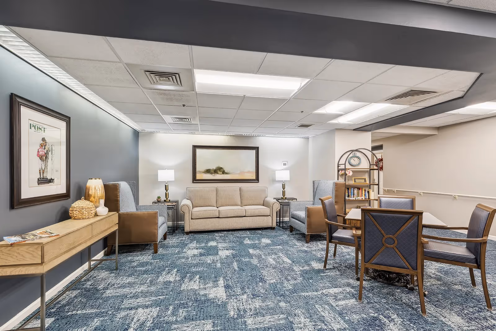 Well-lit senior living common room with a sofa, armchairs, table and chairs, artwork, and a bookshelf.