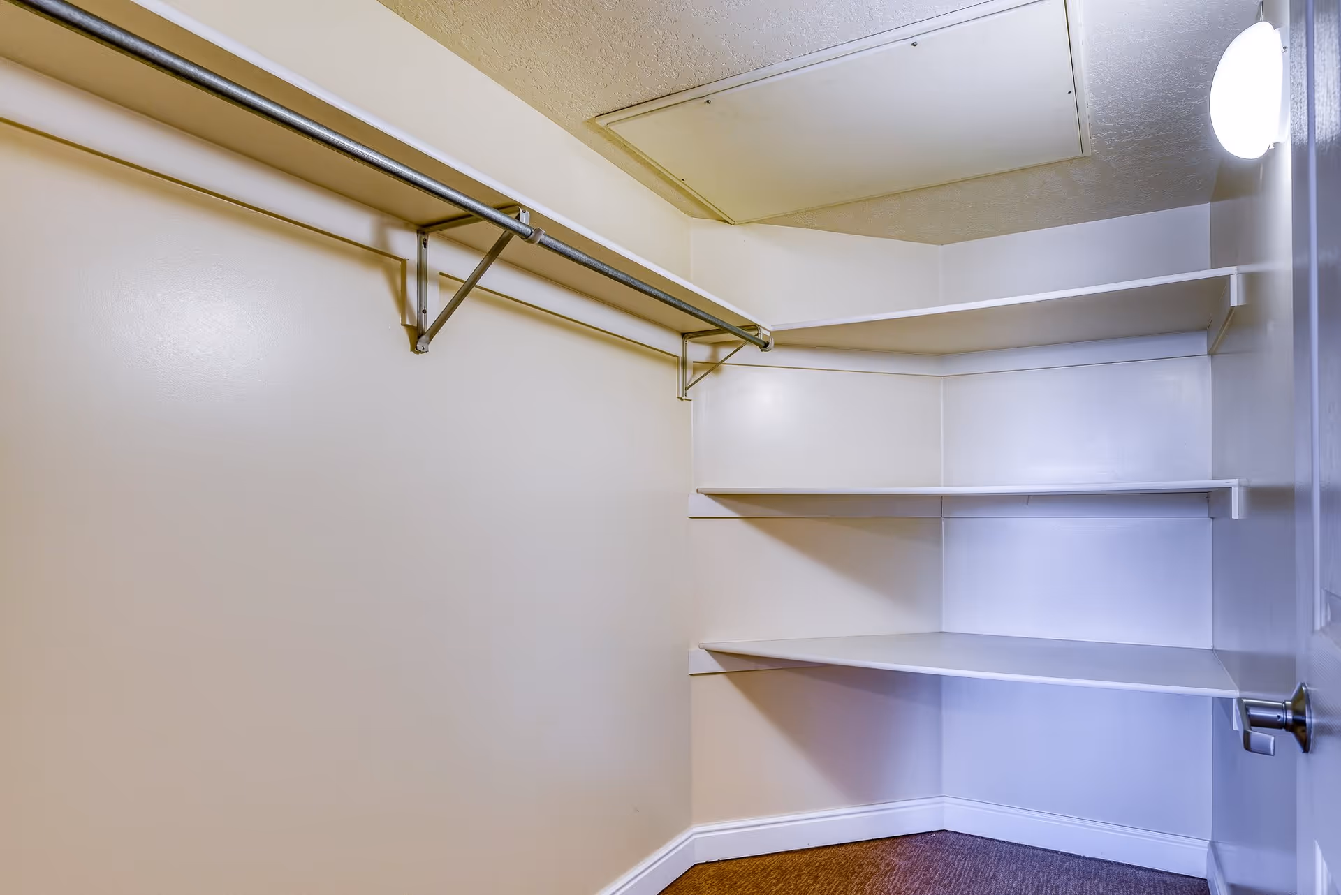 Empty walk-in closet with beige walls, a metal hanging rod, white corner shelves, and a ceiling access panel, illuminated by a wall-mounted light near the door.