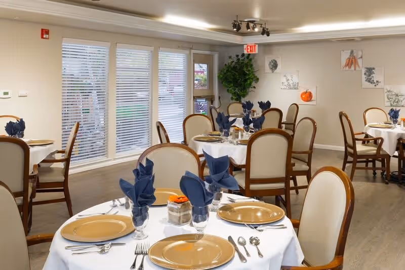 A dining room with several round tables covered with white tablecloths, each set with beige plates, silverware, and glasses with folded dark blue napkins. The room has wooden chairs with cream-colored cushions, large windows with blinds, and light-colored walls decorated with framed botanical prints. There is a green plant near the exit door.