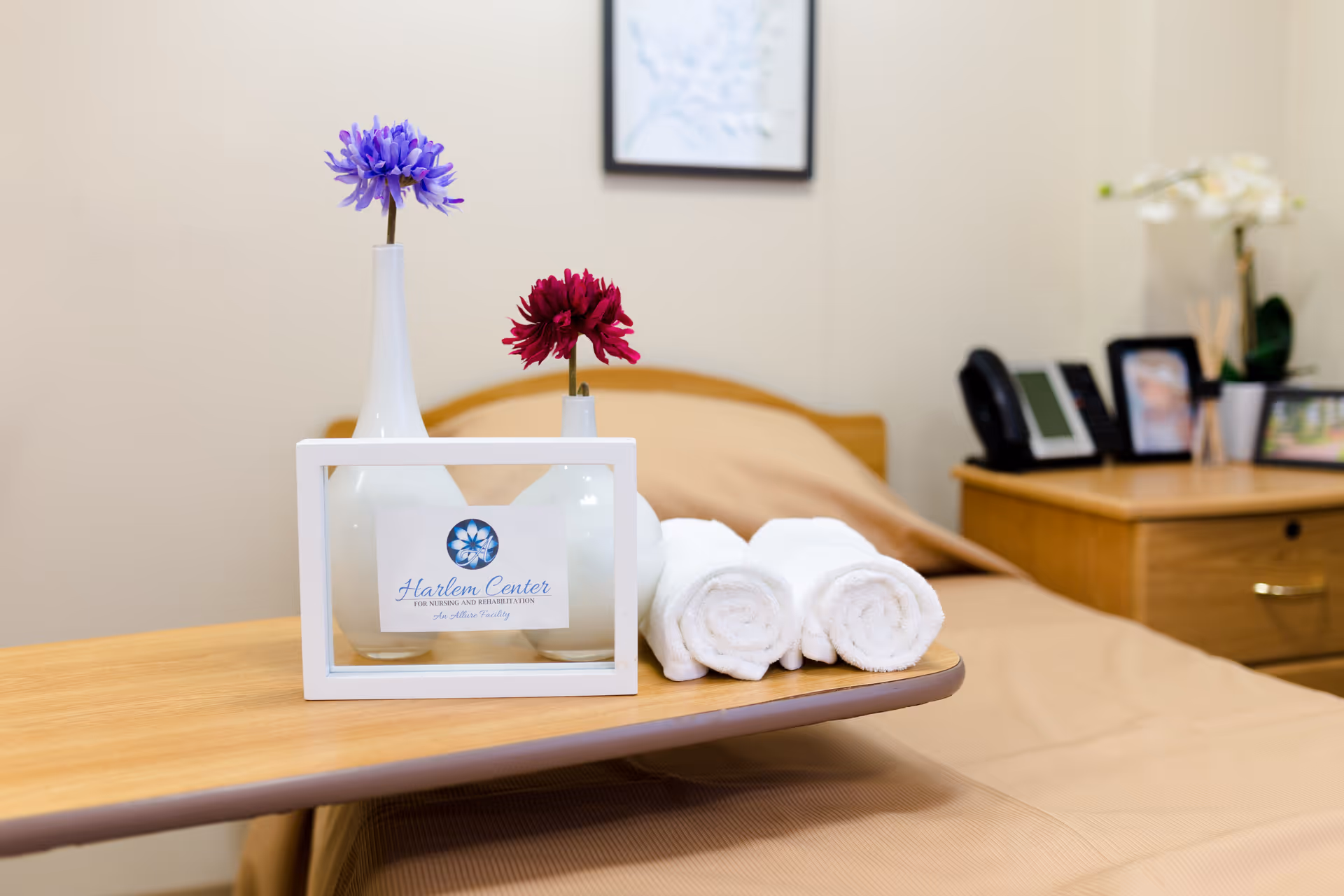 An overbed table on a made bed holding vases with flowers, three rolled towels, and a framed Harlem Center sign in a nursing facility room.