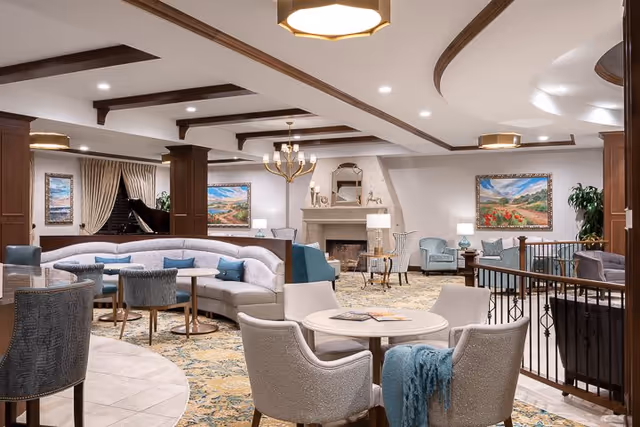 A spacious and elegantly furnished living room area with a curved sofa, multiple armchairs, round tables, and decorative paintings on the walls. The room features a fireplace, ceiling beams, recessed lighting, and a chandelier, creating a warm and inviting atmosphere.